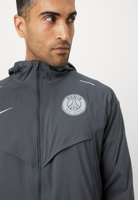 Chaqueta deportiva gris con capucha, con cierre de cremallera, que presenta un parche con logo y acentos reflectantes. Fabricada en un material ligero y texturizado.