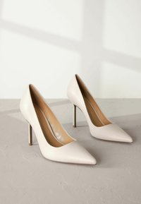 Beige korkokenkä, jossa on terävä kärki, valmistettu sileästä nahasta, ja jossa on tyylikäs muotoilu sekä ohut stiletto-korko, sijoitettuna vaaleanharmaalle pinnalle.