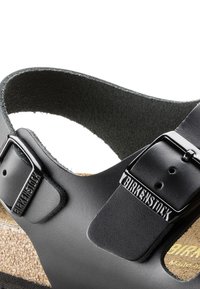 Svart syntetisk sandal med korkfotbädd, justerbar rem med en metallspänne och präglad logotypdetalj. Slät yta.