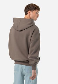 Felpa marrone con una vestibilità rilassata, cappuccio ampio e polsini a costine. Il tessuto sembra morbido con un design minimalista, abbinato a jeans azzurri chiari.