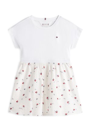 Haut en coton blanc à manches courtes, avec une jupe crème à motifs ornée de petites fleurs rouges et d'accents bleus. Comprend un écusson avec logo sur la poitrine.