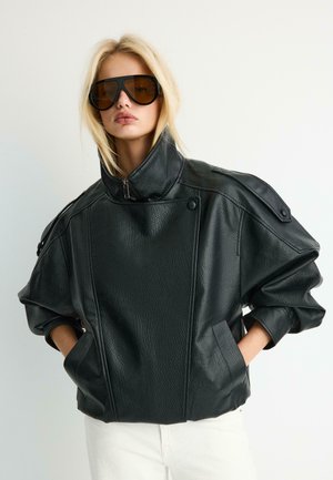 Blondynka w oversize’owej czarnej skórzanej kurtce z wysokim kołnierzem i dużych brązowych okularach przeciwsłonecznych, ręce w kieszeniach kurtki.