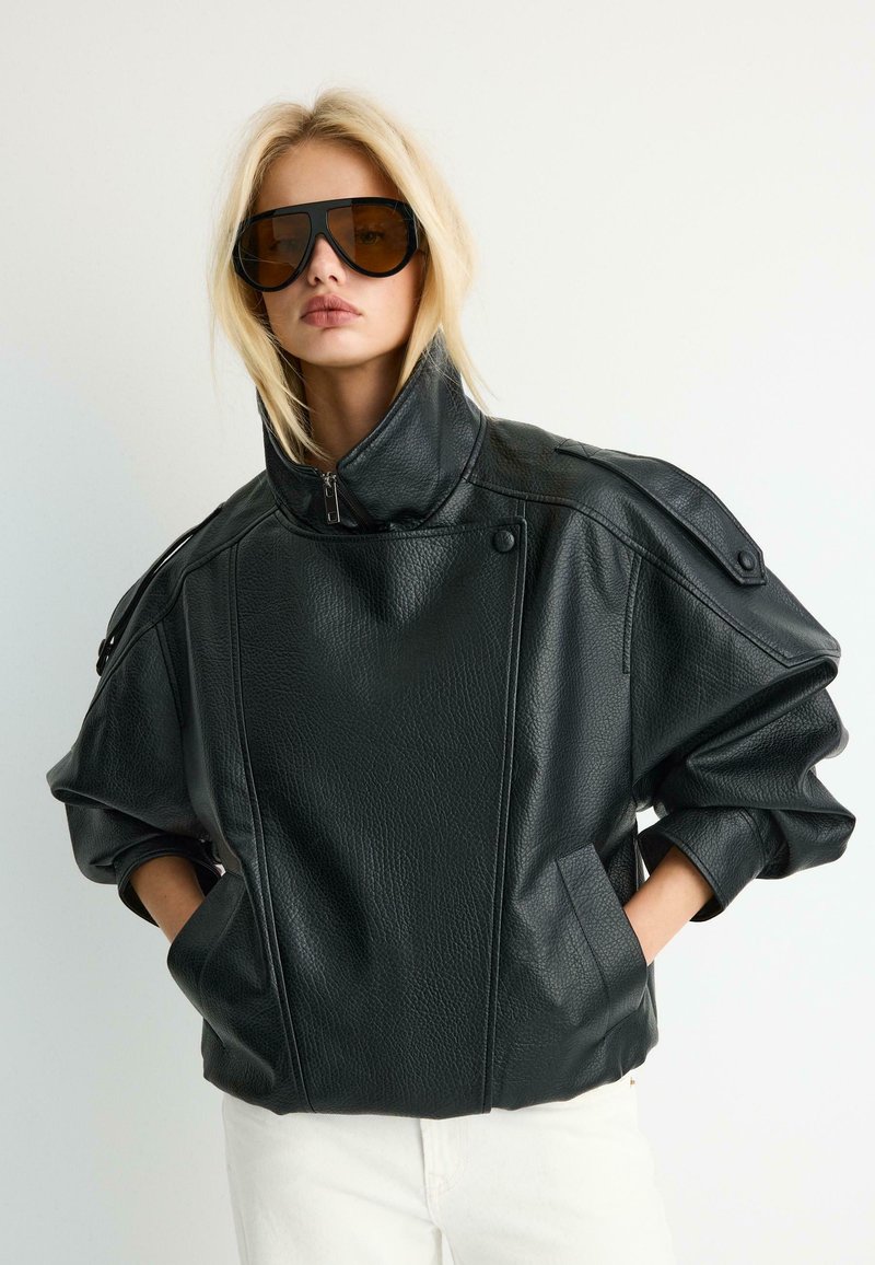 Blonde woman wearing oversized black leather jacket with high collar and large brown-tinted sunglasses, hands in jacket pockets.