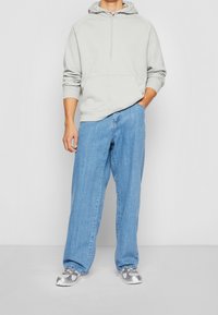 Harmaa huppari, jossa on etutasku, yhdistettynä löysiin sinisiin farkkuihin. Kengät ovat hopeiset urheilukengät. Rentoa ja mukavaa tyyliä.