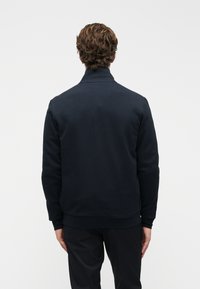 Άνδρας με σγουρά καστανά μαλλιά, φορώντας μαύρο μπουφάν και μαύρο παντελόνι, στέκεται γυρισμένος σε μια απλή λευκή φόντο.