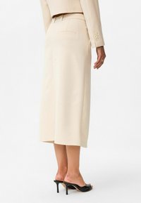 Beige midi-rok met een op maat ontworpen snit, zijzakken en een gladde textuur. Gecombineerd met slanke zwarte hoge hakken.