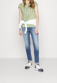 T-shirt vert clair avec un logo script, pull blanc noué à la taille, jean bleu, et baskets colorées avec un mélange d'accents bleus, jaunes et roses.