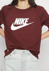 Burgunderfärgad sweatshirt med en vit Nike-logotyp. Har rundad halsringning och raglanärmar. Tyget verkar mjukt och bekvämt.