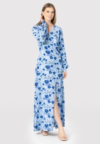 Maxi kjole i lyseblå stof med blåt blomsterprint, med lange ærmer, V-udskæring og en frontslids. Båret med nude hæle.