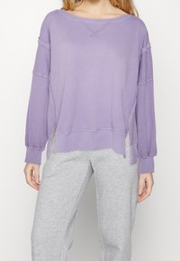 Felpa viola oversize con maniche lunghe, spalle scese, polsini a costine e spacchi laterali. Texture liscia e vestibilità casual.