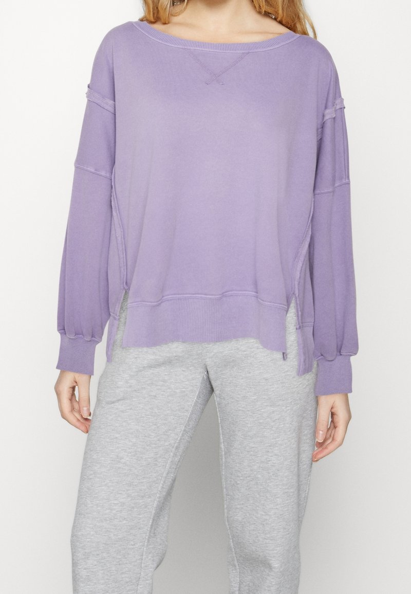 Felpa viola oversize con maniche lunghe, spalle scese, polsini a costine e spacchi laterali. Texture liscia e vestibilità casual.