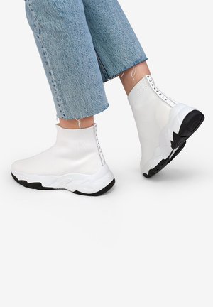 Zapatillas estilo calcetín blancas con parte superior de punto acanalado, suela negra contrastante y cremalleras laterales con un detalle de marca vertical.