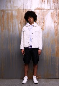 Giacca di jeans bianca sopra una felpa leggera tie-dye blu, abbinata a pantaloni cargo neri e sneakers bianche. Posizionata davanti a uno sfondo in metallo arrugginito.