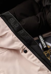 Jasnoróżowa podszewka kurtki z czarną podszewką, z markowym zamkiem błyskawicznym i czarnym napem. Widoczna fakturowana tkanina oraz detal z logo.