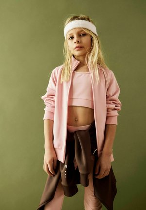 Young girl in pink sportswear and white headband stands with hands down against plain olive background.