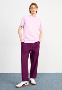 T-shirt in cotone rosa chiaro con una piccola grafica e maniche corte, abbinata a pantaloni larghi di un viola scuro e sneakers bianche con dettagli neri.