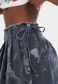 Hochtaillierter, dunkler Denim-Rock mit einer gebundenen Taillen-Schnur, Knopfverschluss, Falten und bedruckten Designs, darunter Herzen und Sterne, auf einer Person, die ihr Hemd hochhebt.