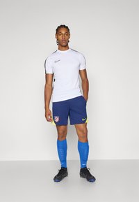 Hvid atletisk T-shirt med sorte accenter, og navysorte shorts med gult kant og et logo, blå sokker og sorte fodboldstøvler.