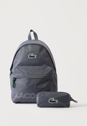 Γκρι σακίδιο Lacoste και αντίστοιχη κασετίνα με πράσινο λογότυπο κροκόδειλου σε λευκό φόντο.