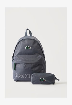 Γκρι σακίδιο Lacoste και αντίστοιχη κασετίνα με πράσινο λογότυπο κροκόδειλου σε λευκό φόντο.
