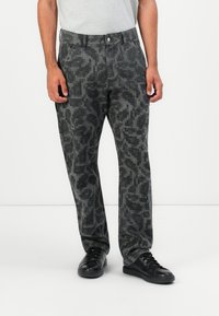 Mann som har på seg svart og grå bukser med pixelmønster, hvit t-skjorte tucked in, og svarte joggesko, står mot en ensfarget lys bakgrunn.