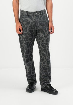 Mann som har på seg svart og grå bukser med pixelmønster, hvit t-skjorte tucked in, og svarte joggesko, står mot en ensfarget lys bakgrunn.