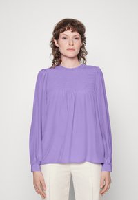 Blusa lavanda com mangas compridas, textura franzida no decote, e um colarinho redondo. O tecido suave cai solto até à metade do quadril.