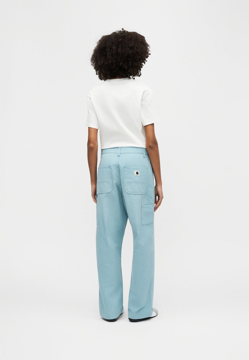 Personne aux cheveux bouclés portant une chemise blanche à manches courtes et un pantalon large bleu clair, debout de dos sur un fond uni.
