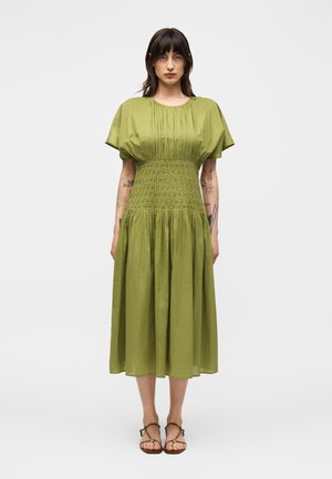 Mujer con cabello oscuro que lleva un vestido midi verde con mangas abullonadas y cintura fruncida, de pie con los brazos relajados, mostrando tatuajes en los brazos.