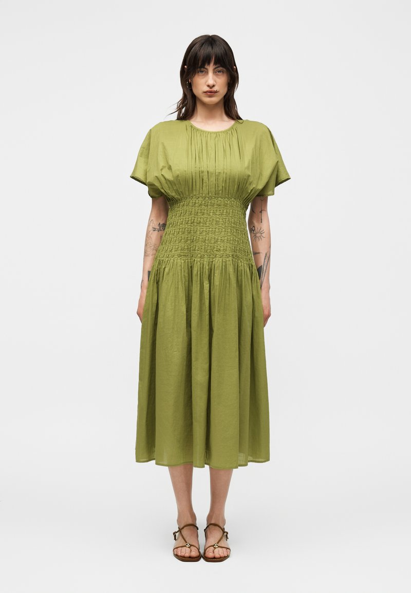 Γυναίκα με σκούρα μαλλιά που φοράει πράσινο μίντι φόρεμα με φουσκωτά μανίκια και σφηκοφωλιά στη μέση, στέκεται με χαλαρά χέρια, δείχνοντας τατουάζ στα χέρια.