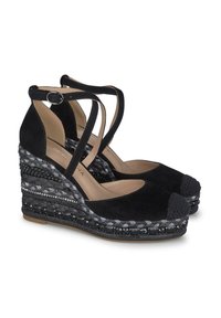 Zwarte suède wedge sandalen met kruislings gespen rond de enkel, ronde neus en gevlochten textuur op de hak, voorzien van metalen accenten.