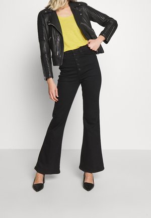 Giacca da motociclista in pelle nera con dettagli a coste, top giallo, jeans neri a vita alta a zampa d'elefante e tacchi neri con punta affilata.