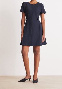 Marinblå kortärmad klänning i texturerad tyg; har en åtsittande midja och en lätt utsvängd kjol med två dekorativa knappar vid midjan.
