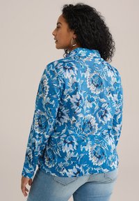 Blå blommig blus med långa ärmar, som har stora, överlappande blommor i nyanser av blått och vitt. Mjuk tyg med en strukturerad krage.