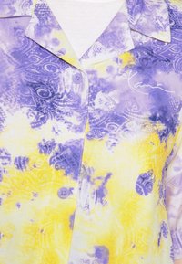 Camicia a maniche corte con motivi tie-dye gialli e viola, chiusura a bottoni e un sottile dettaglio con firma bianca sul davanti.