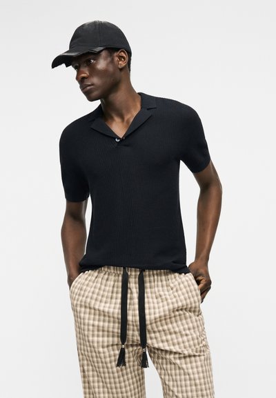 Homme portant une chemise noire à manches courtes avec col, une casquette noire et un pantalon beige à carreaux avec des cordons noirs, debout les mains dans les poches.