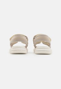 Beige sandaler med teksturerte stropper og spenne-lukkinger. Polstret hvit såle med preget merkenavn. Bakfra vises stroppes design.