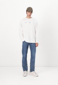 White long-sleeve shirt with blue text, blue jeans, and white sneakers. The model wears a green cap, standing against a plain white background.