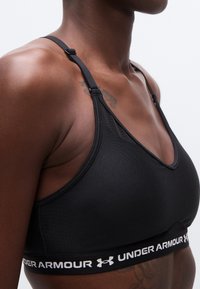 Κοντινό πλάνο ενός ατόμου που φοράει μαύρο αθλητικό σουτιέν Under Armour με ρυθμιζόμενους ιμάντες και λωρίδα με λογότυπο, εμφανίζοντας τον ώμο, το στήθος και το άνω μέρος του κορμού.
