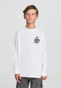 Biała bawełniana koszulka z długim rękawem, z okrągłym dekoltem, z czarnym graficznym logo na piersi i luźnym krojem.