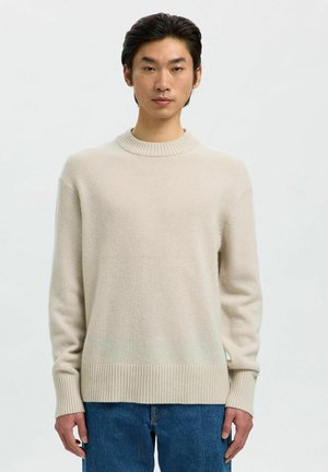 Joven con cabello oscuro que lleva un suéter de cuello redondo de canalé beige y jeans azules frente a un fondo claro y sencillo.