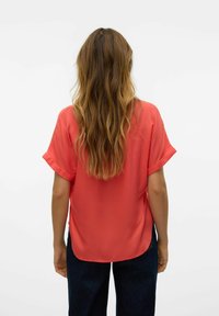 Blusa corallo con maniche corte arrotolate, realizzata in tessuto liscio, caratterizzata da una vestibilità ampia e un orlo curvo. Indossata con pantaloni blu scuro.