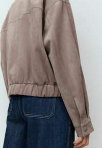 Taupe suède jack met ribbelzoom, paneeldetails en met knopen sluitende manchetten. Gecombineerd met donkerblauwe denim. Gelaagd stof en gestructureerde vorm zichtbaar.