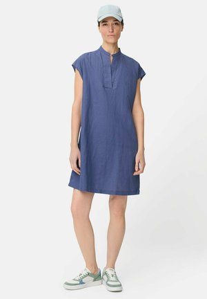 Kvinde iført en blå kortærmet kjole, lyseblå baseballkasket og hvide sneakers med grønne og blå detaljer, stående mod en ensfarvet baggrund.