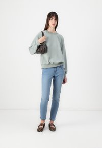 Felpa verde chiaro, jeans slim-fit blu e ballerine con stampa leopardata. Il modello tiene una piccola borsa marrone. Colori solidi e vestibilità casual.