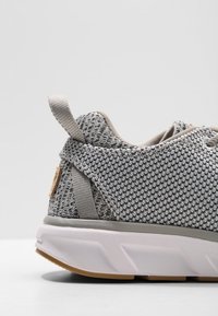 Chaussure de sport tricotée grise avec une tige texturée, un tirant à boucle au talon et une semelle blanche rembourrée. Présente des accents marron minimalistes.