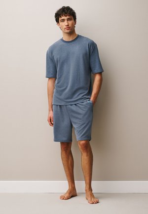 Junger Mann mit lockigem Haar steht barfuß da und trägt ein passendes blaues kurzärmeliges Hemd und Shorts vor einer beigen Wand.