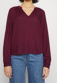 Blusa bordeaux a maniche lunghe con scollatura a V, caratterizzata da spalle arricciate e una vestibilità comoda, abbinata a jeans a gamba larga blu chiaro.