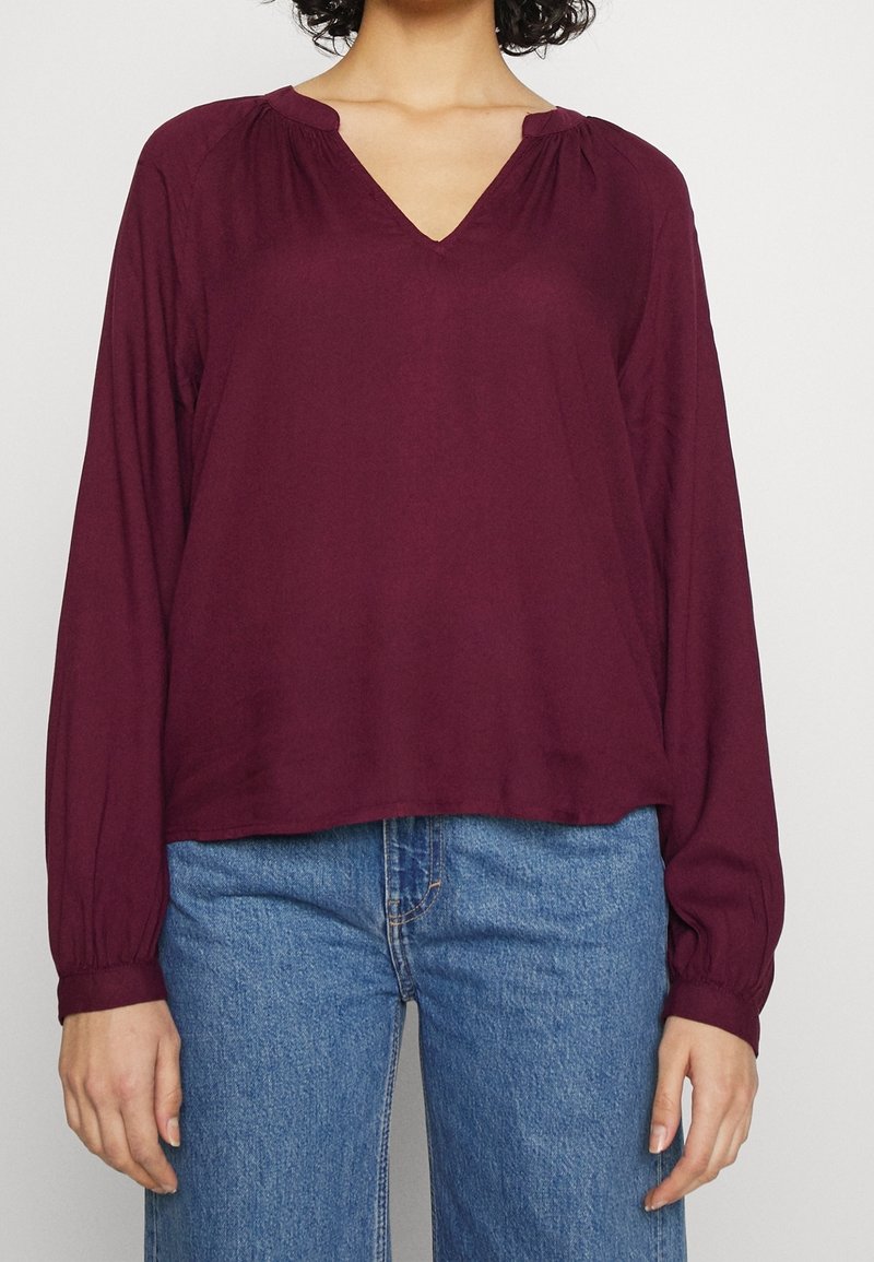 Blusa bordeaux a maniche lunghe con scollatura a V, caratterizzata da spalle arricciate e una vestibilità comoda, abbinata a jeans a gamba larga blu chiaro.