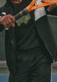 Person som bär svarta sportkläder med en grön krokodil-logotyp, håller en orange tennisracket i mittslag på en bana.